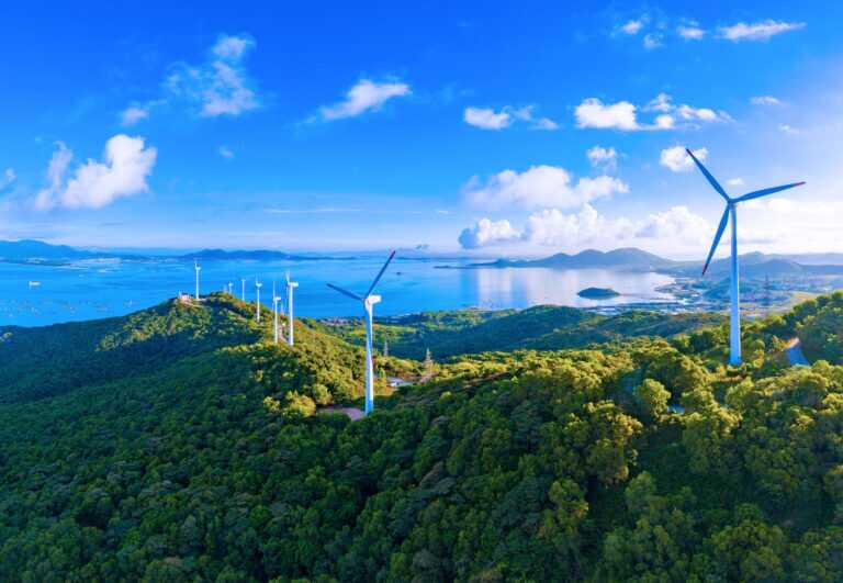 a wind farm featuring multiple white wind turbines spread across green forested hills with a blue sea and islands in the background.