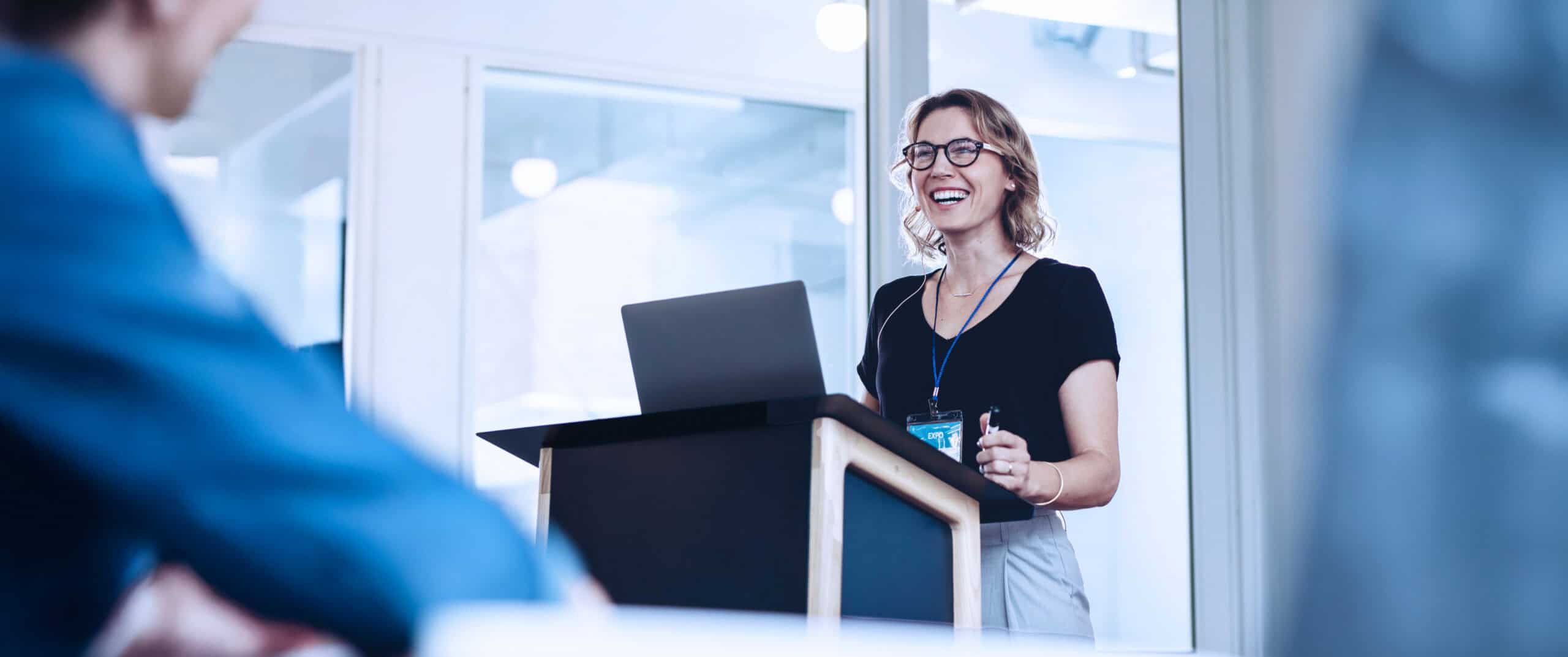 Photograph showing a woman standing behind a podium with a laptop, giving a presentation in a modern office setting.
