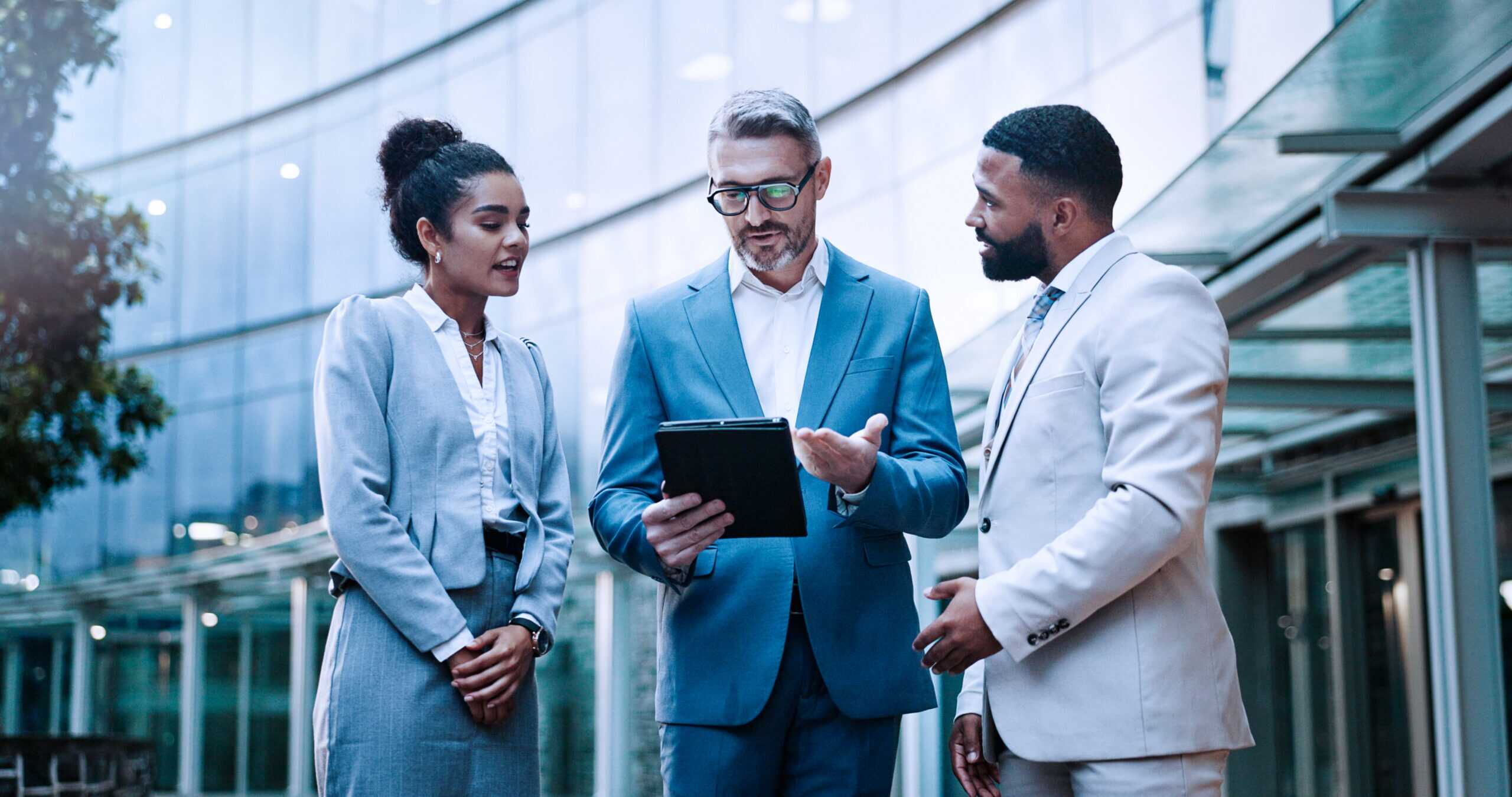 Photograph of three business professionals standing outdoors in front of a modern glass building, engaged in discussion while one person holds a tablet.