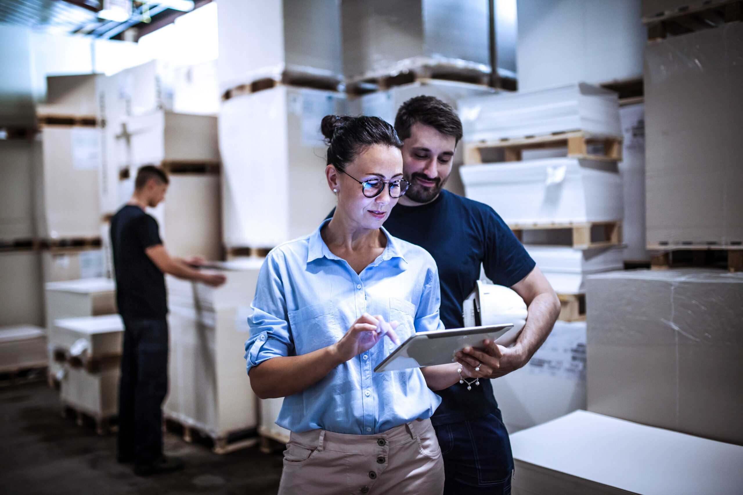 Two warehouse workers using a digital tablet for inventory management, with stacks of large cardboard boxes and pallets in background.