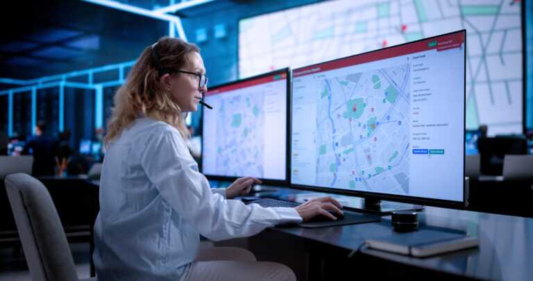 Photograph of a person wearing a headset working at a desk with two large monitors displaying detailed maps with various coloured markers and data points.