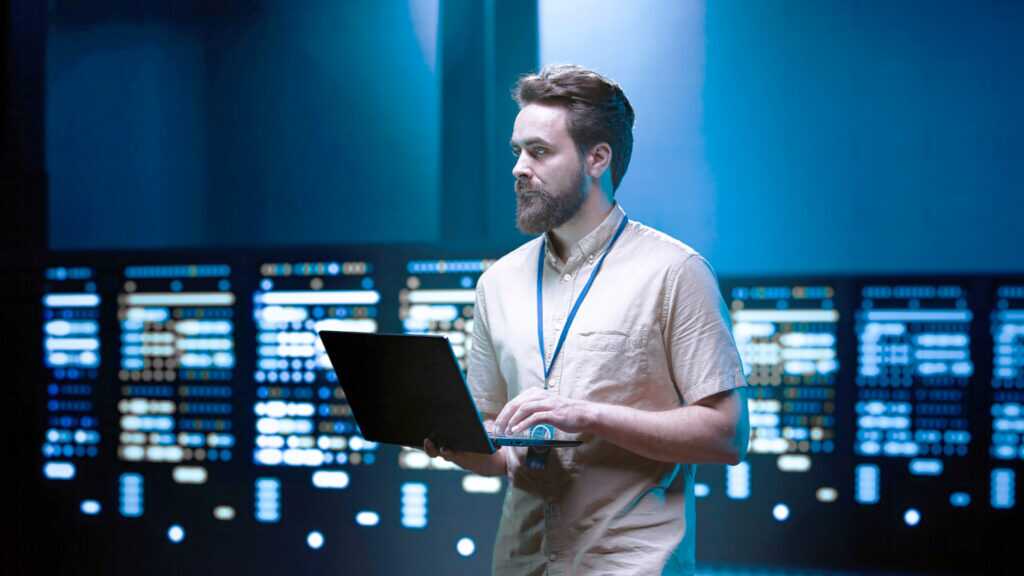 A person standing in a server room holding and working on a laptop, surrounded by racks of illuminated servers.