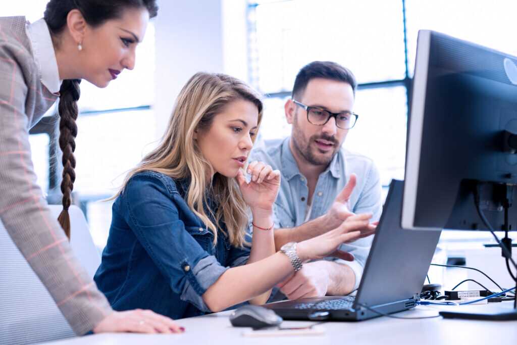 Photograph showing three colleagues collaborating around a desk with a laptop and desktop monitor in a bright office setting.