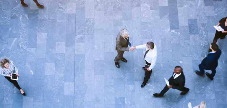 Aerial photograph showing seven business professionals walking and interacting on a large grey tiled floor. The two central figures are shaking hands.