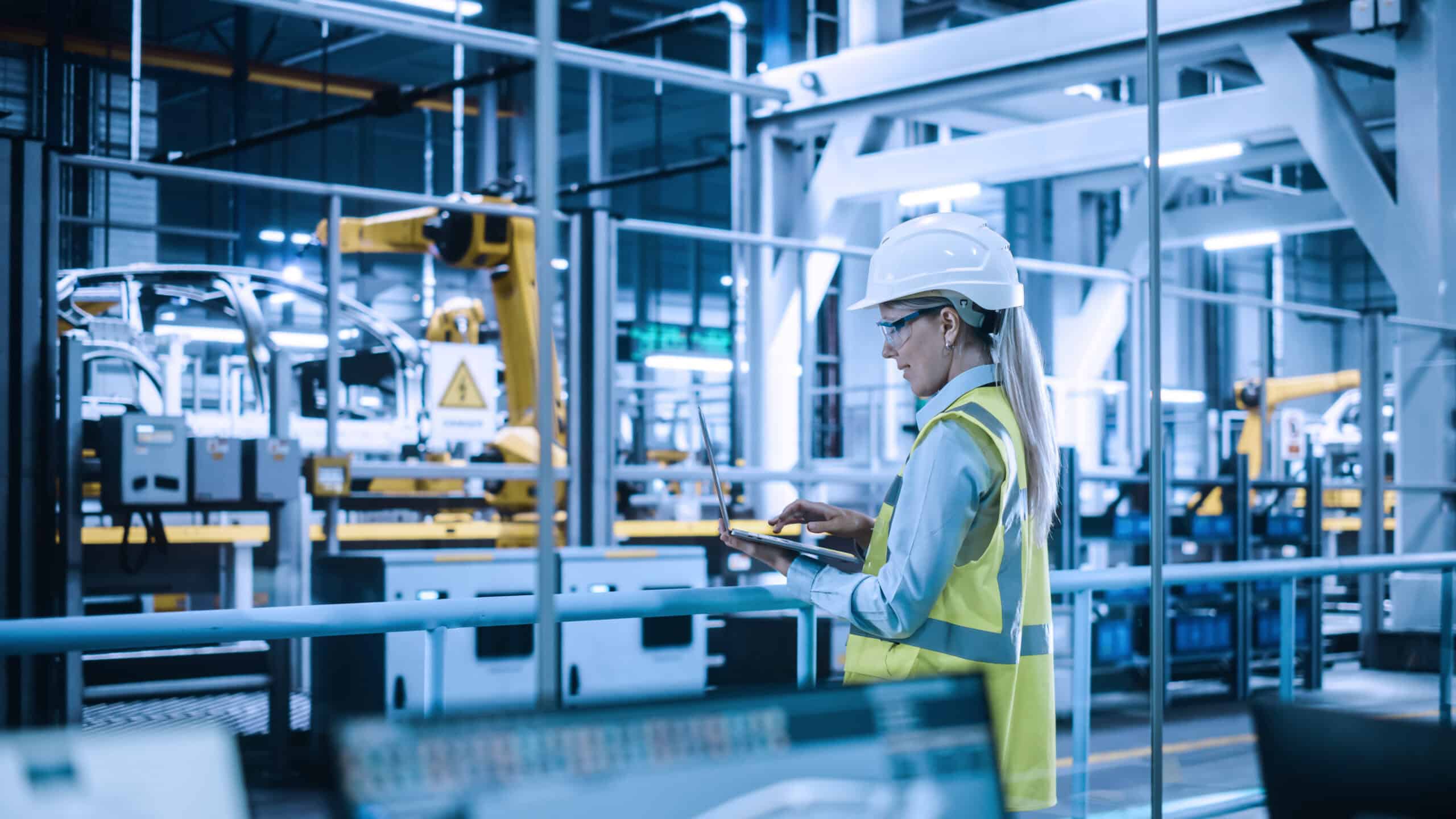 Photograph of a factory worker wearing a white hard hat and yellow safety vest using a tablet in an industrial setting.
