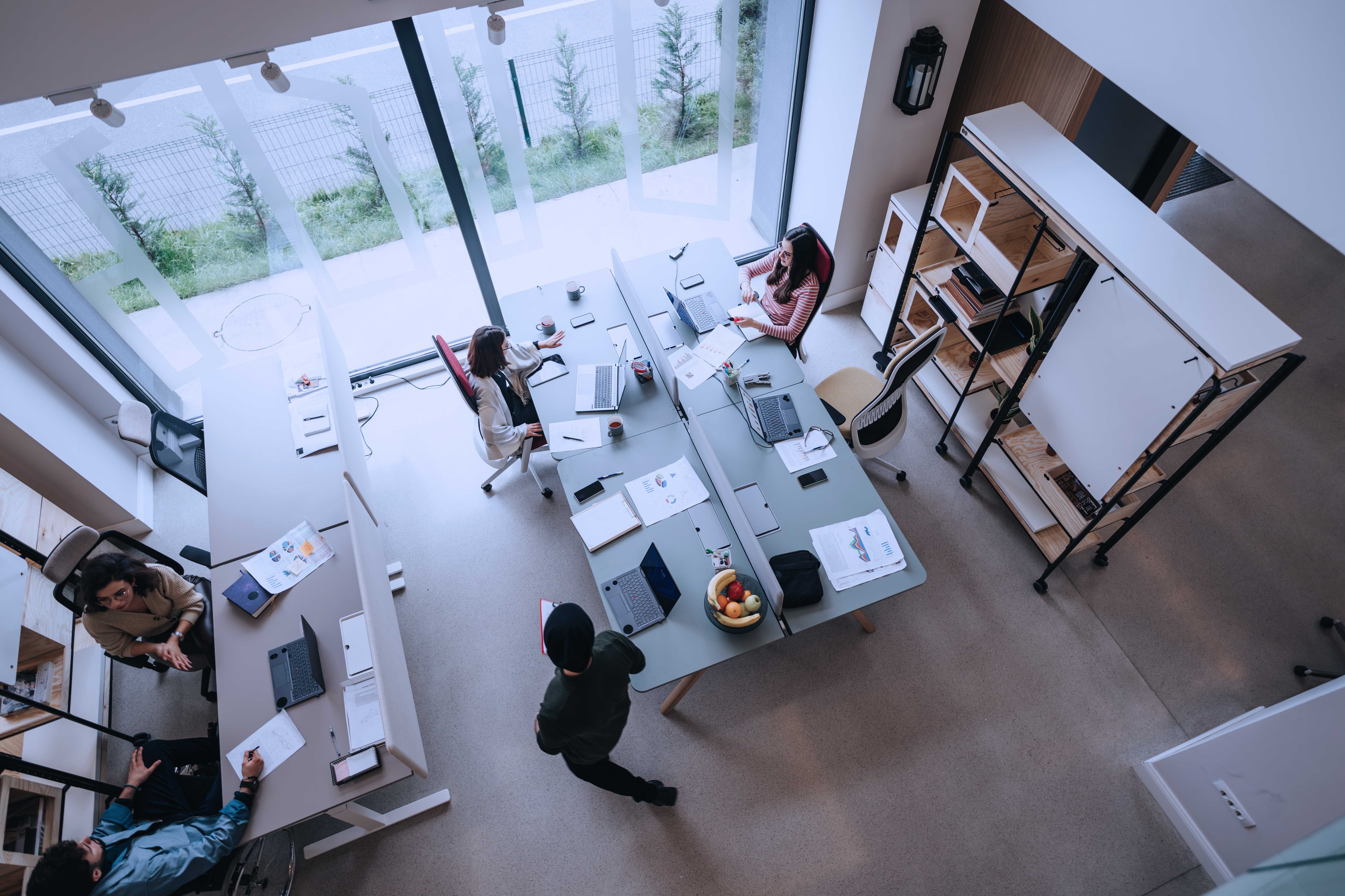 Photograph showing a modern office space with four people engaged in work around two desks arranged in an L-shape near large windows.