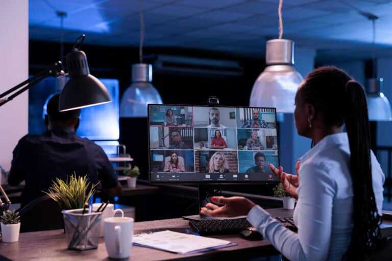 Photograph of a women in a modern office participating in a video conference call displayed on a computer monitor with nine participants in a grid layout.