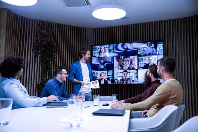 Business meeting with five people seated around a conference table and one person standing, presenting to a large screen displaying a video call with multiple participants.