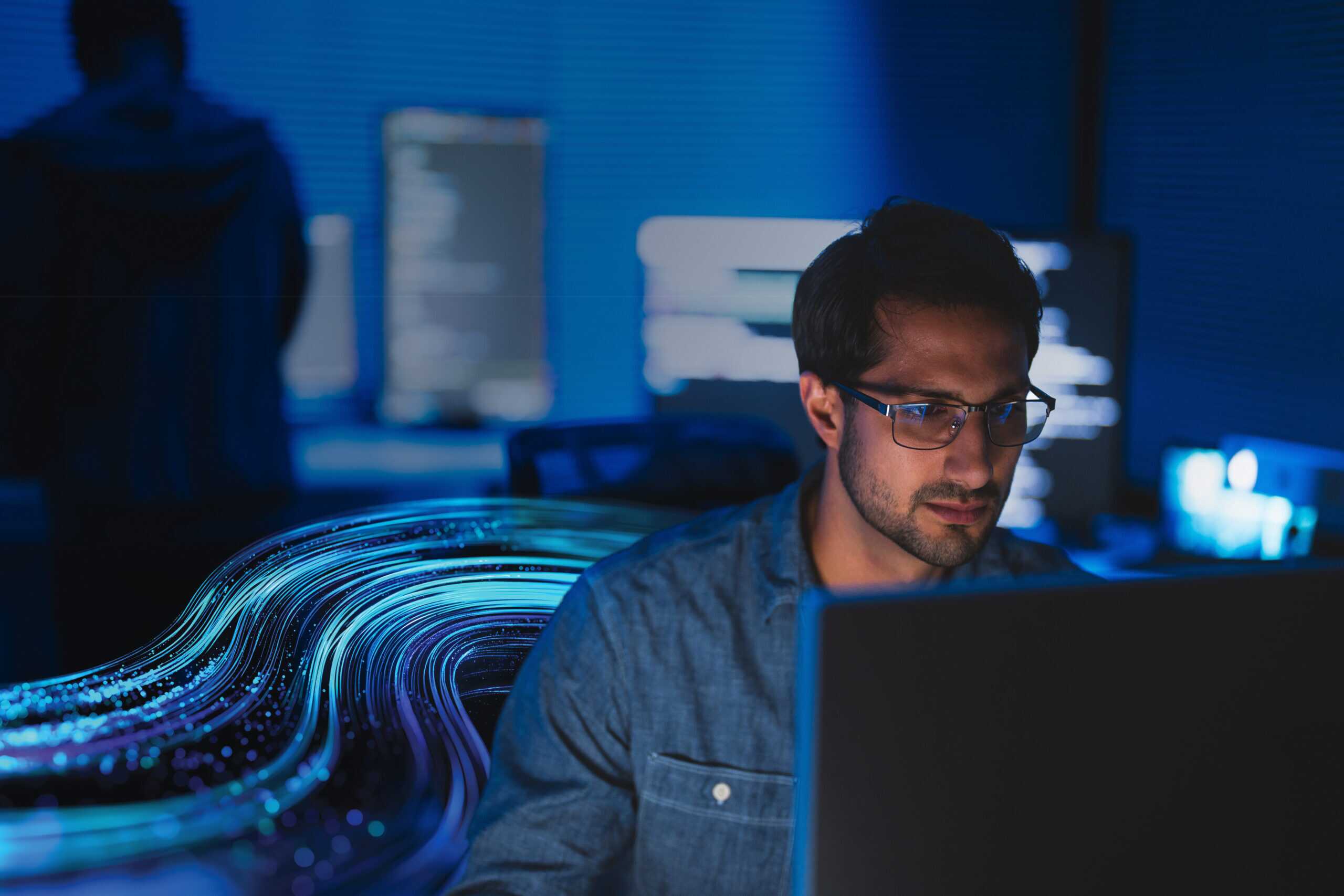 Person working on a laptop in a dark room with multiple illuminated computer screens in the background. The SCC wave graphic flows from behind his right shoulder.