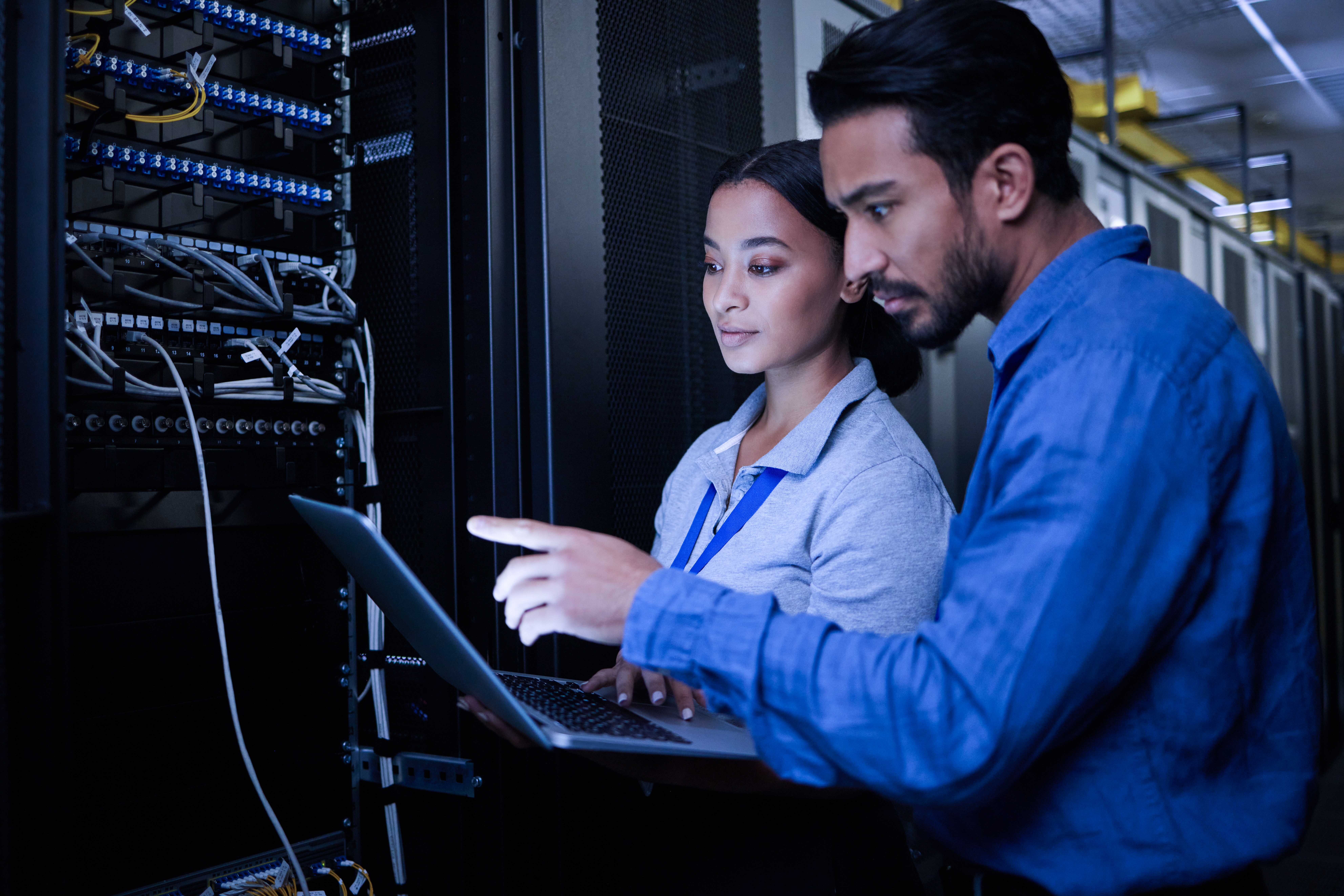 Two IT professionals working in a server room, with one person holding a laptop and the other pointing at the screen.