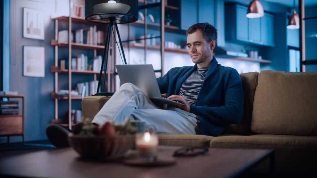 Photograph of a person sitting on a sofa using a laptop in a dimly lit living room with bookshelves and modern decor.