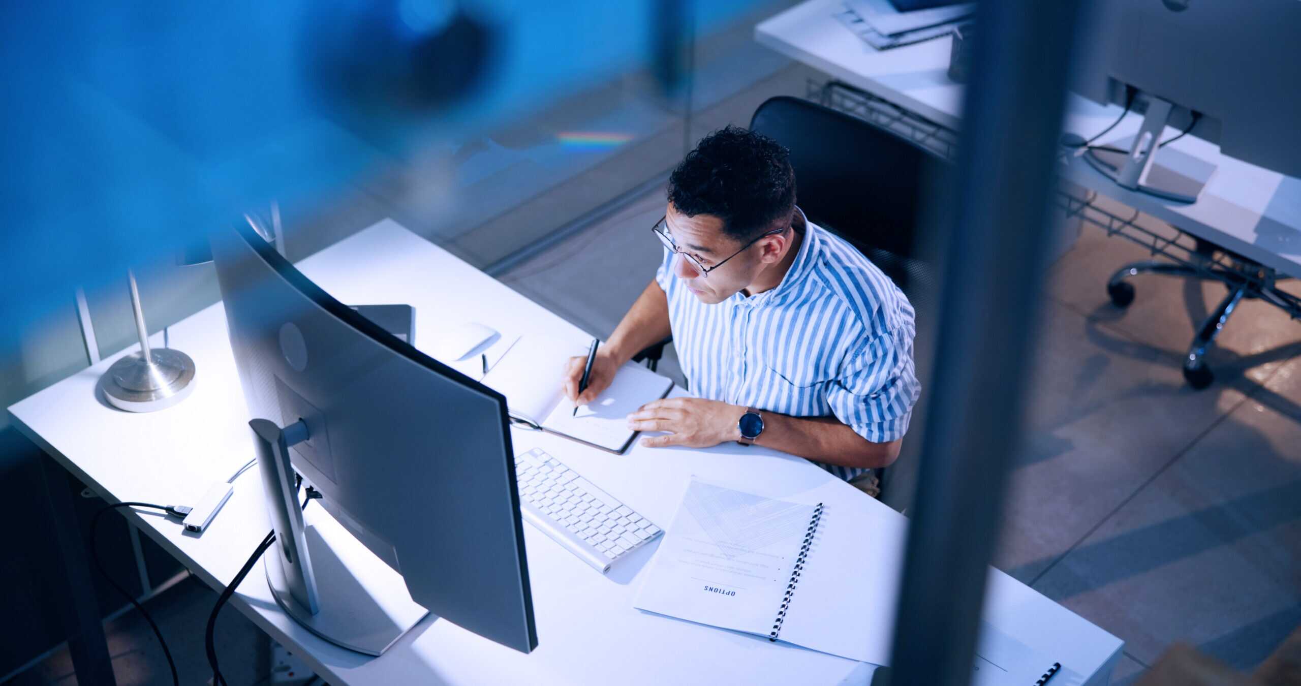 Photograph of a person working at a desk with dual monitors, writing in a notebook surrounded by documents and office supplies.
