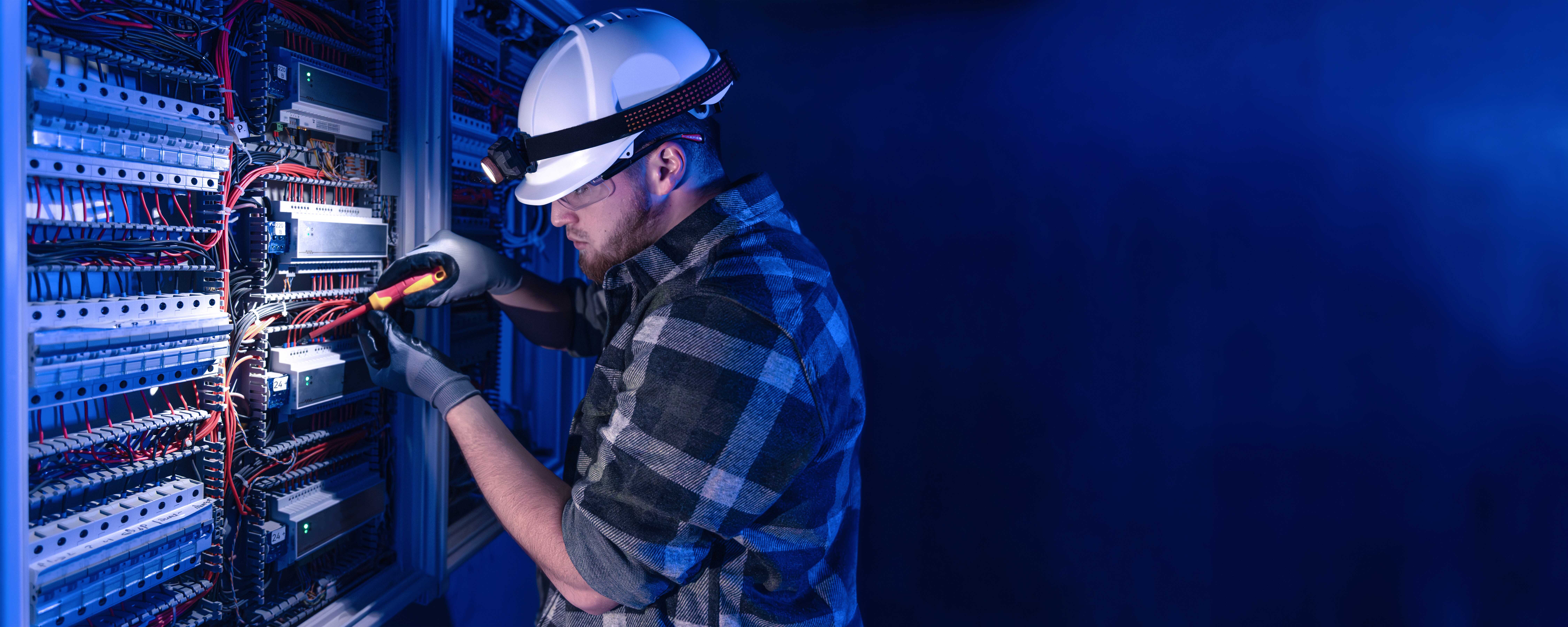 Photograph of a technician wearing a hard hat and gloves working on an electrical panel with numerous wires and circuit breakers.