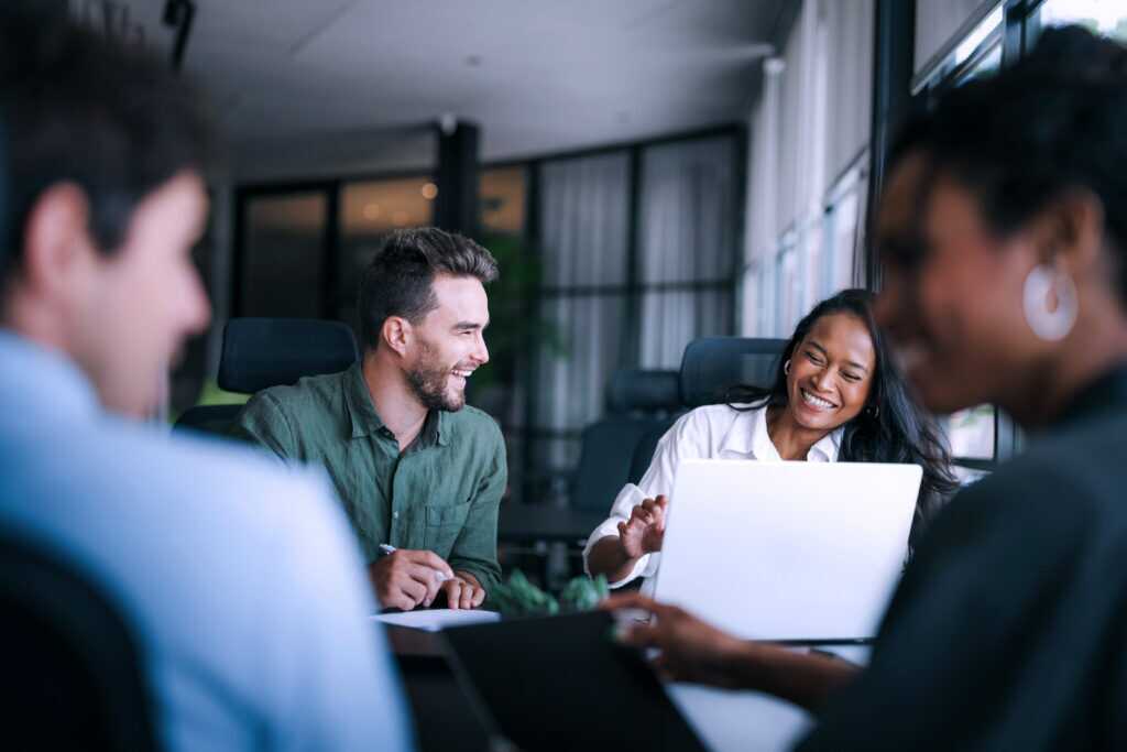 Photograph of a group meeting in a modern office with four people engaged in discussion around a table. Focus is on two individuals, one holding a laptop and explaining something.