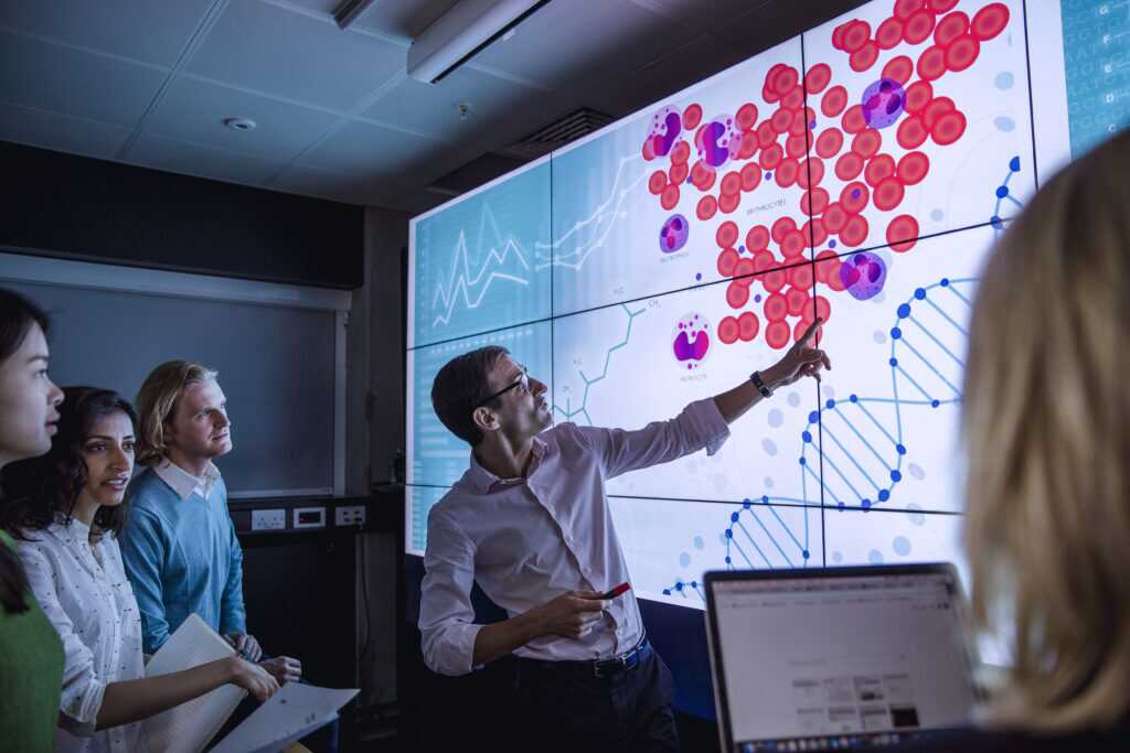 Photograph showing a group of people in a presentation setting with a large digital screen displaying scientific data.