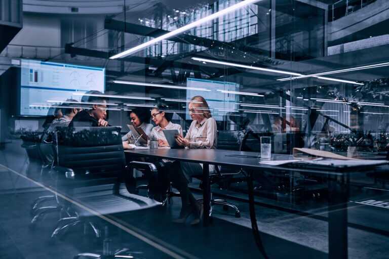 Photograph of a business meeting in a modern office with six professionals seated around a conference table, discussing documents and digital tablets.