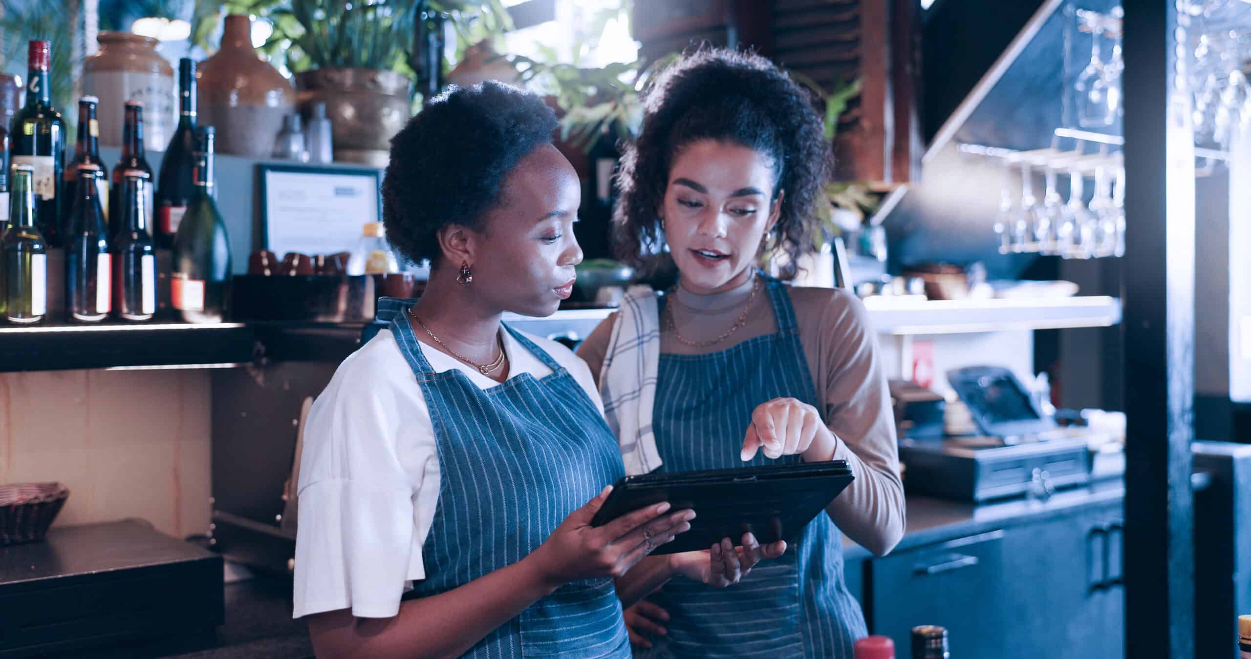Photograph of two bar staff wearing blue aprons standing behind a bar counter, looking at and interacting with a digital tablet.
