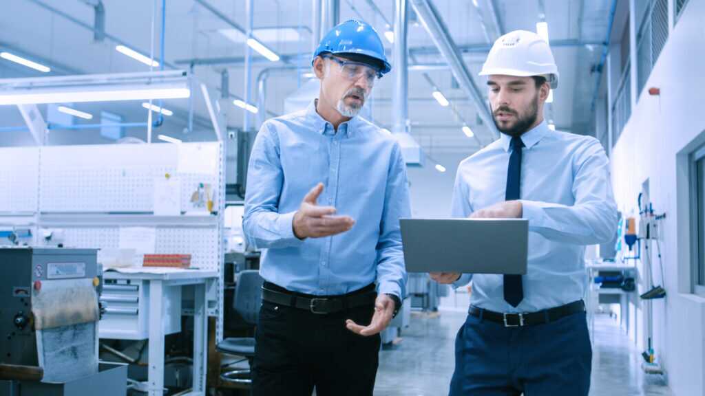 Photograph showing two engineers in a factory setting, both wearing safety helmets and discussing work while one holds a laptop.