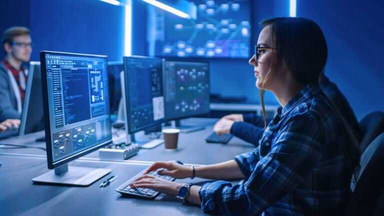 Photograph showing a person working on multiple computer monitors displaying code and data visualisations.