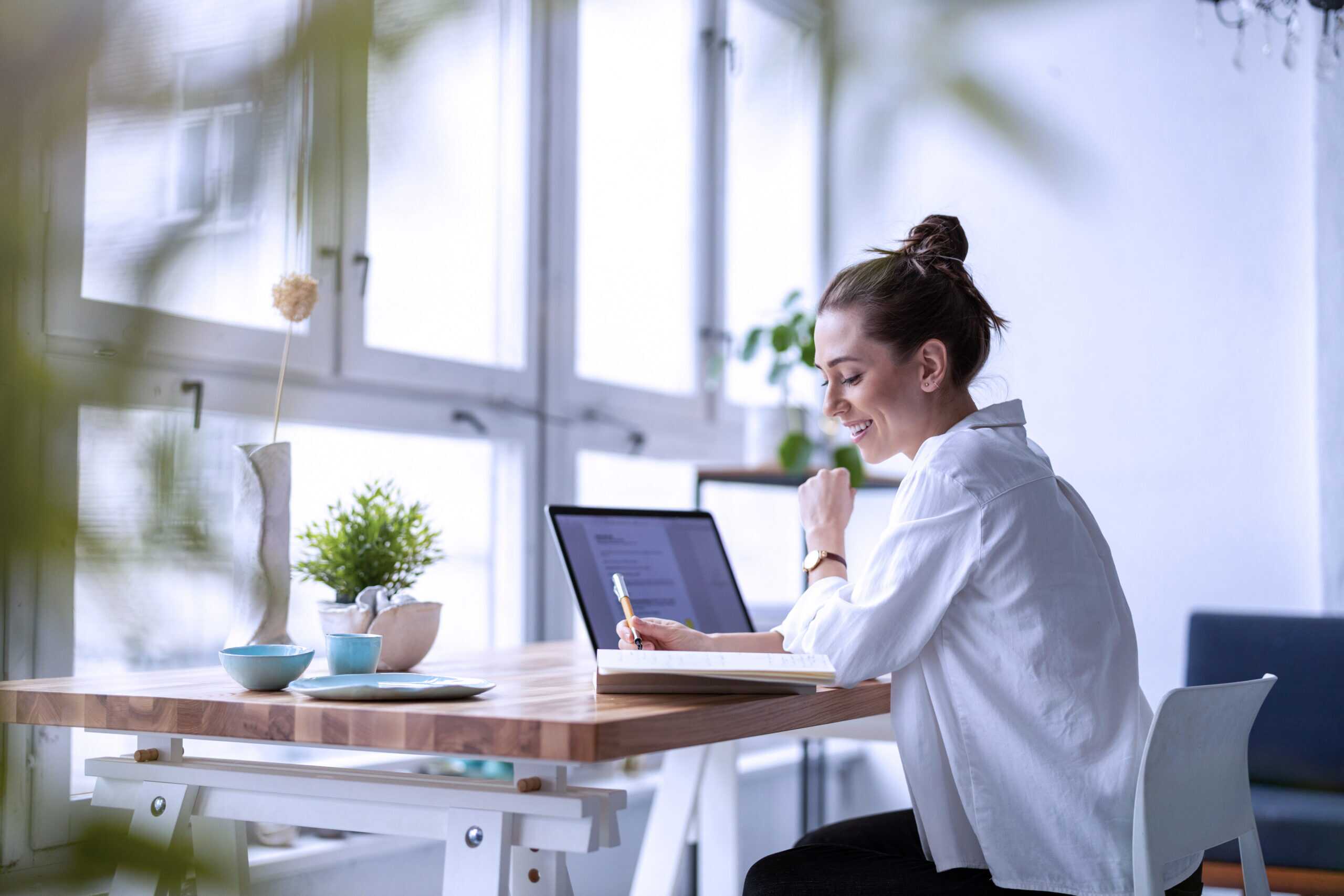 Photograph of a person sitting at a wooden desk in a bright room, working on a laptop and writing in a notebook.