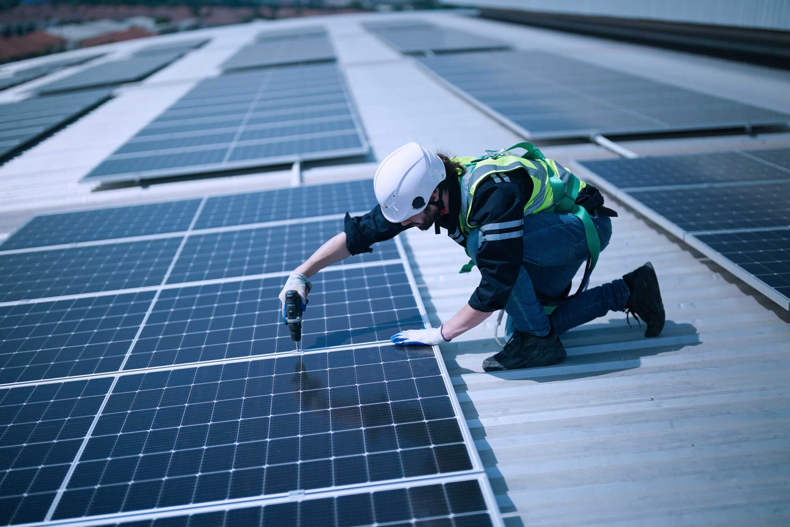 A worker wearing a white helmet and green safety vest installing or inspecting solar panels on a rooftop.