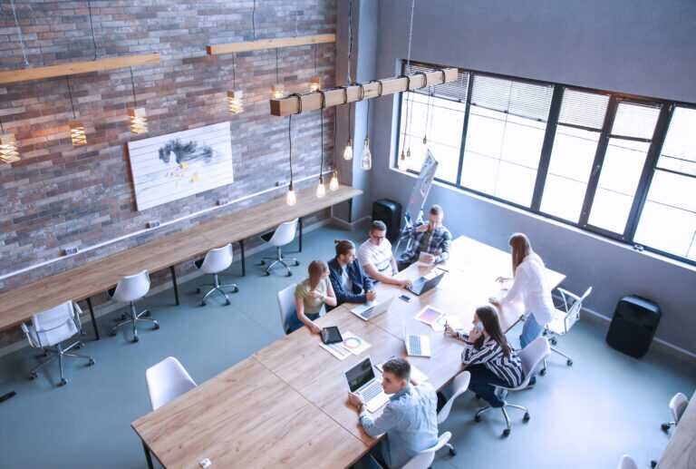 Photograph of a modern office space featuring a group of six people collaborating around a large wooden table with laptops and documents.