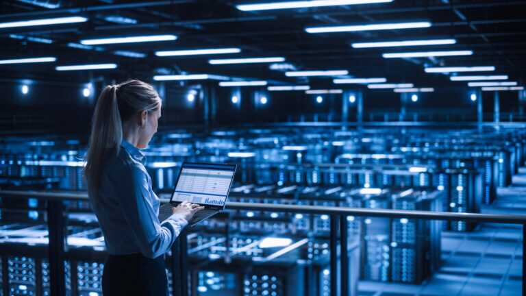 Woman standing in a large, dimly lit data centre filled with rows of server racks illuminated by blue lights.