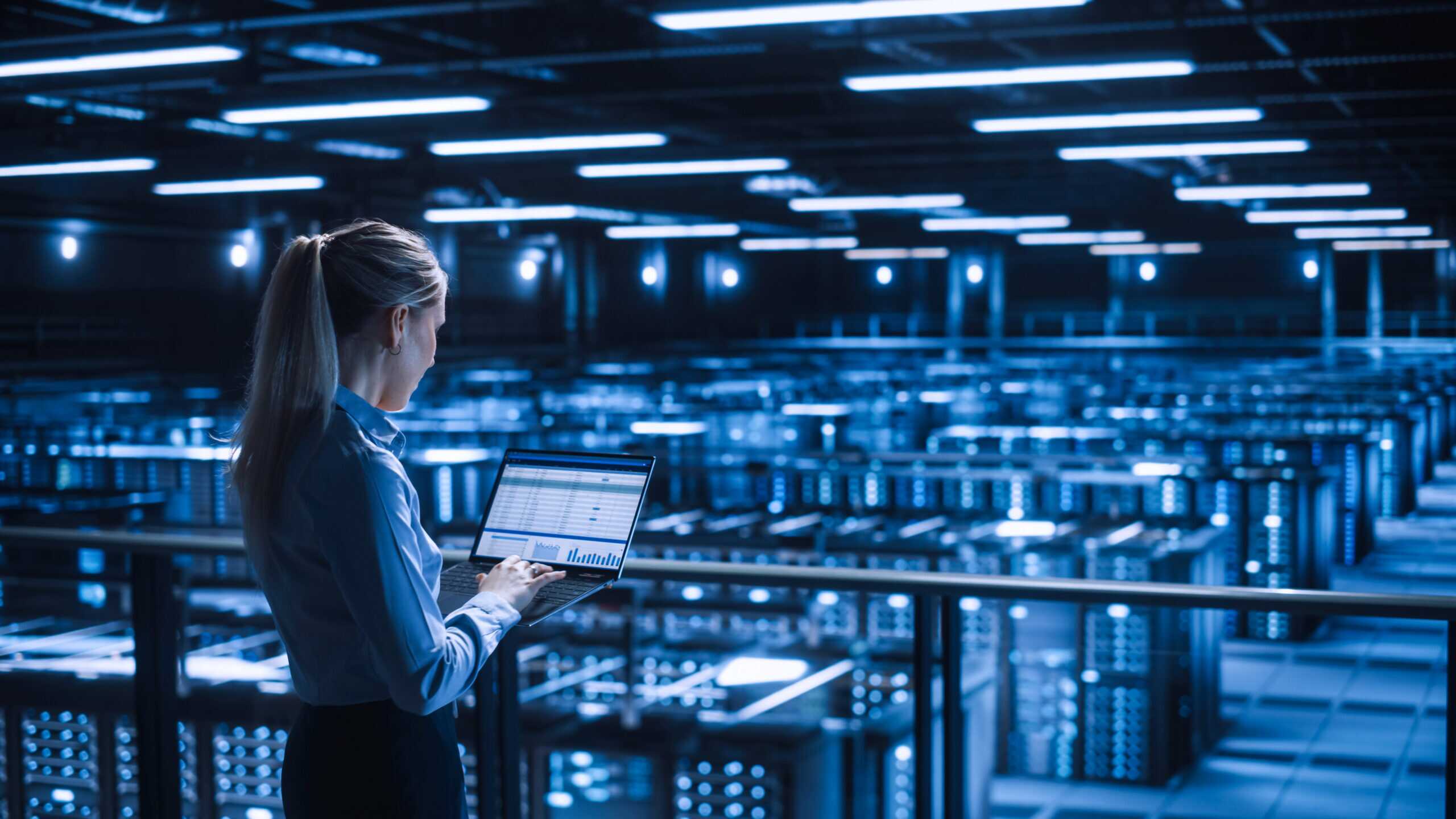 Woman standing in a large, dimly lit data centre filled with rows of server racks illuminated by blue lights.