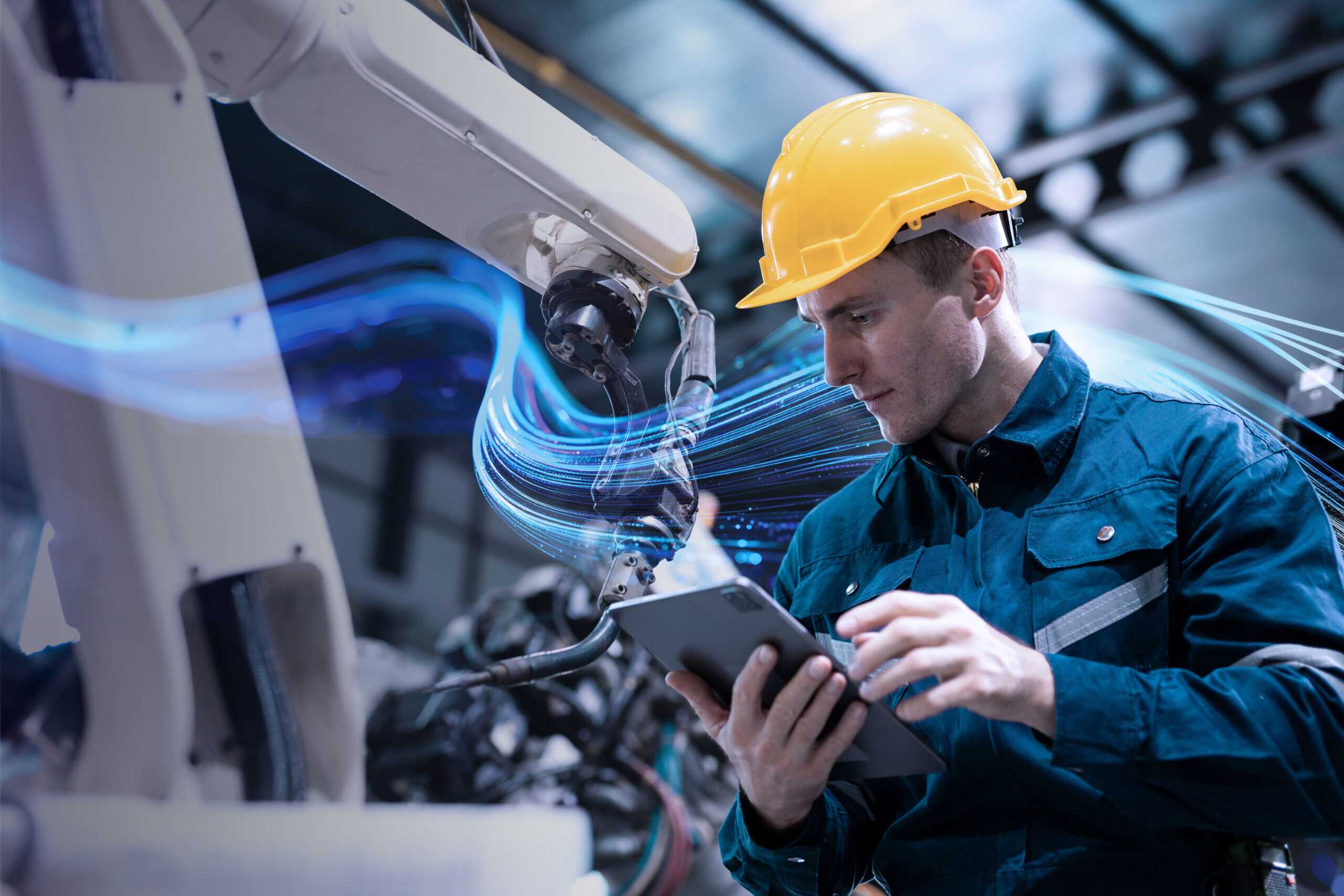 Factory worker wearing a yellow hard hat and blue uniform operating a robotic arm using a tablet, with the SCC wave graphic in the background.